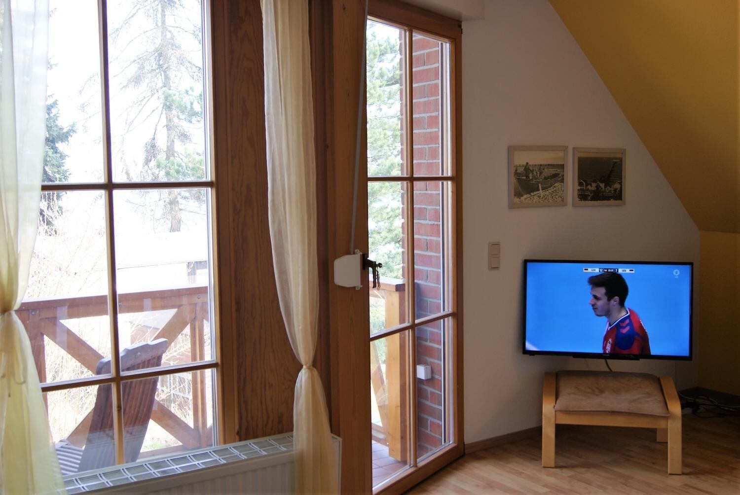 Quarto com televisão em suporte, janelas amplas com cortinas, piso de madeira e parede de tijolos.