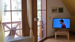 A room with a TV on a stand, large windows with curtains, a wooden floor, and a brick wall. - Blick auf Vilm (Putbus)