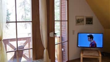 Quarto com televisão em suporte, janelas amplas com cortinas, piso de madeira e parede de tijolos.