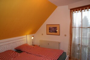 A bedroom with a sloped ceiling, a radiator, a framed picture on the wall, and a window with sheer curtains. - Blick auf Vilm (Putbus)