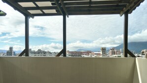 Un espacio al aire libre cubierto con un banco de madera, vista a un paisaje urbano con edificios y montañas al fondo.