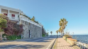A coastal road with a stone building on the left, a paved walkway, and palm trees.