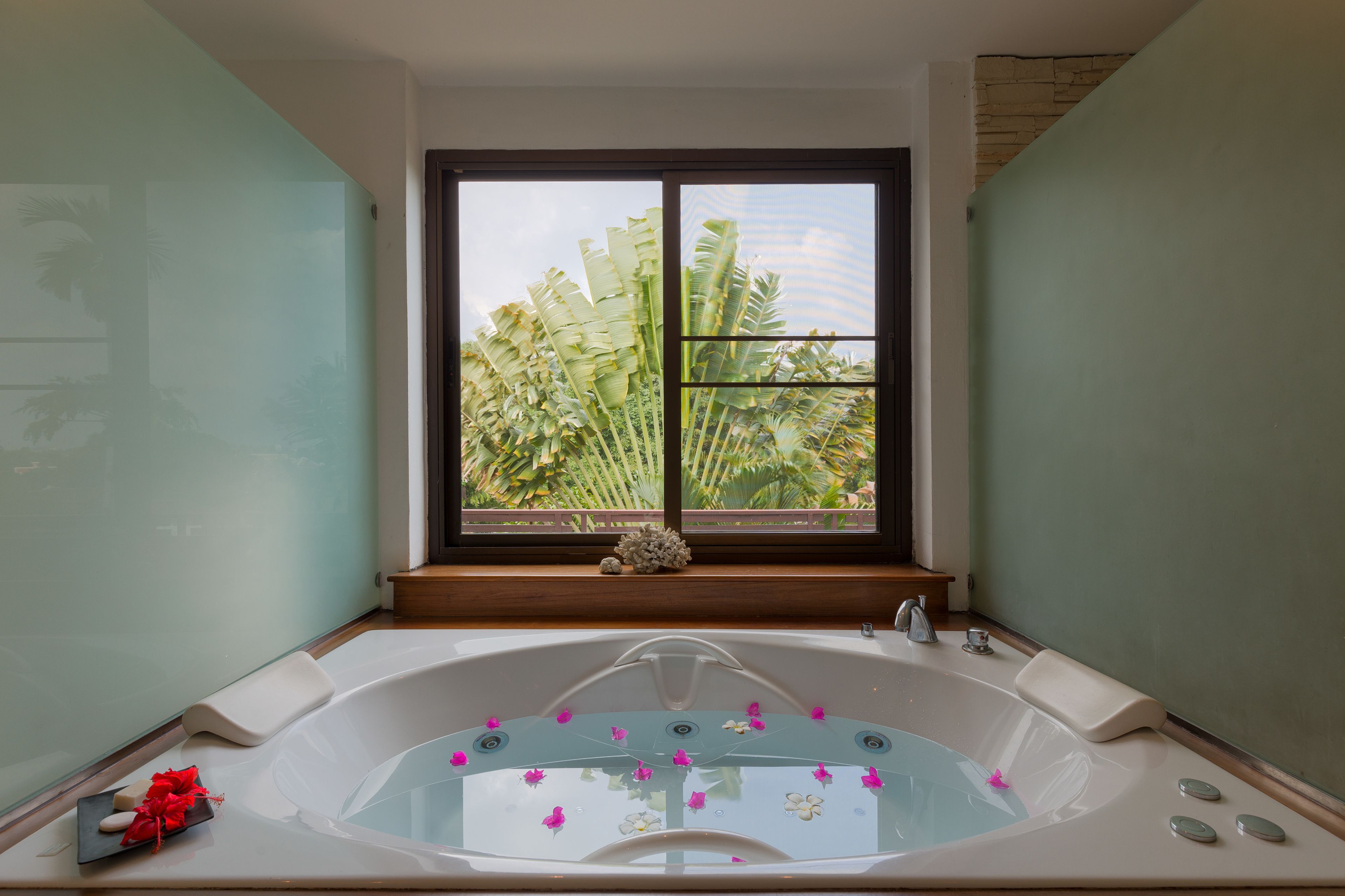 A bathroom with a large bathtub, a window with a view of palm trees, and a small shelf with decorative items.