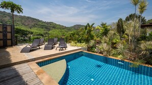 A swimming pool with lounge chairs, surrounded by tropical plants and a view of a hilly landscape. - Villa Oxavia (Rawai)