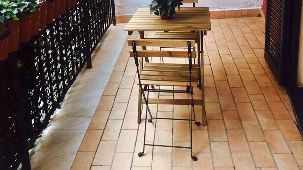 A covered outdoor patio with a wooden table and chairs, a black metal fence, and potted plants. - Il Cuore di Napoli (Naples)