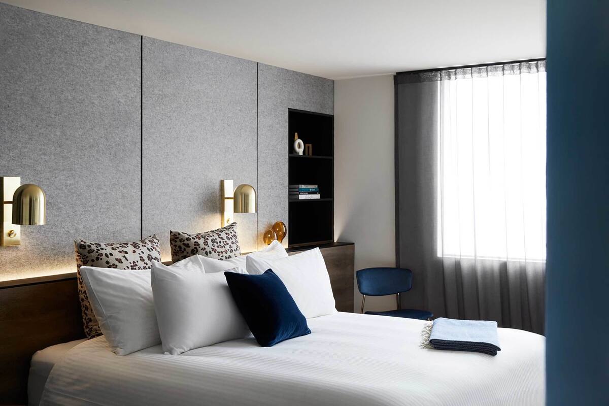 Ett modernt hotellrum med en stor säng, två väggmonterade lampor, ett sidobord, en stol och en bokhylla.