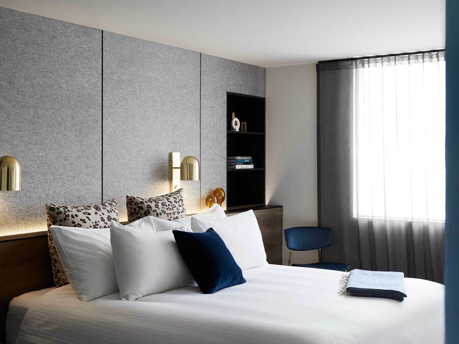 Quarto de hotel moderno com uma cama grande, duas luminárias fixadas na parede, uma mesinha de cabeceira, uma cadeira e uma estante de livros.