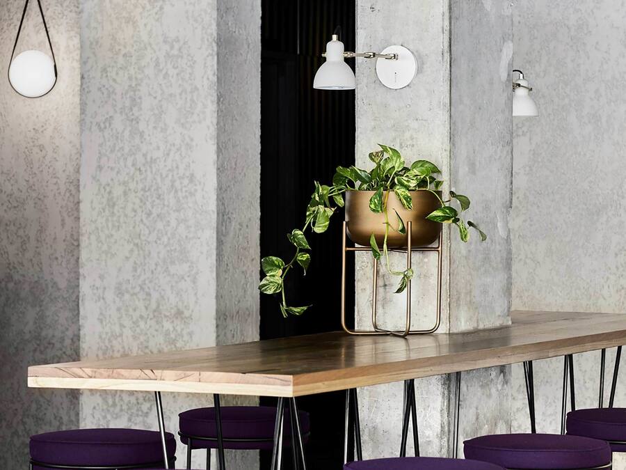 A modern bar area with a long wooden table, purple bar stools, a hanging light fixture, and a potted plant on the wall.