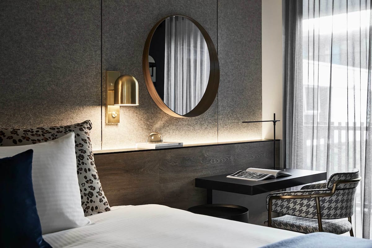 Ett modernt hotellrum med en stor säng, ett skrivbord med en stol, en rund spegel och ett fönster med gardiner.