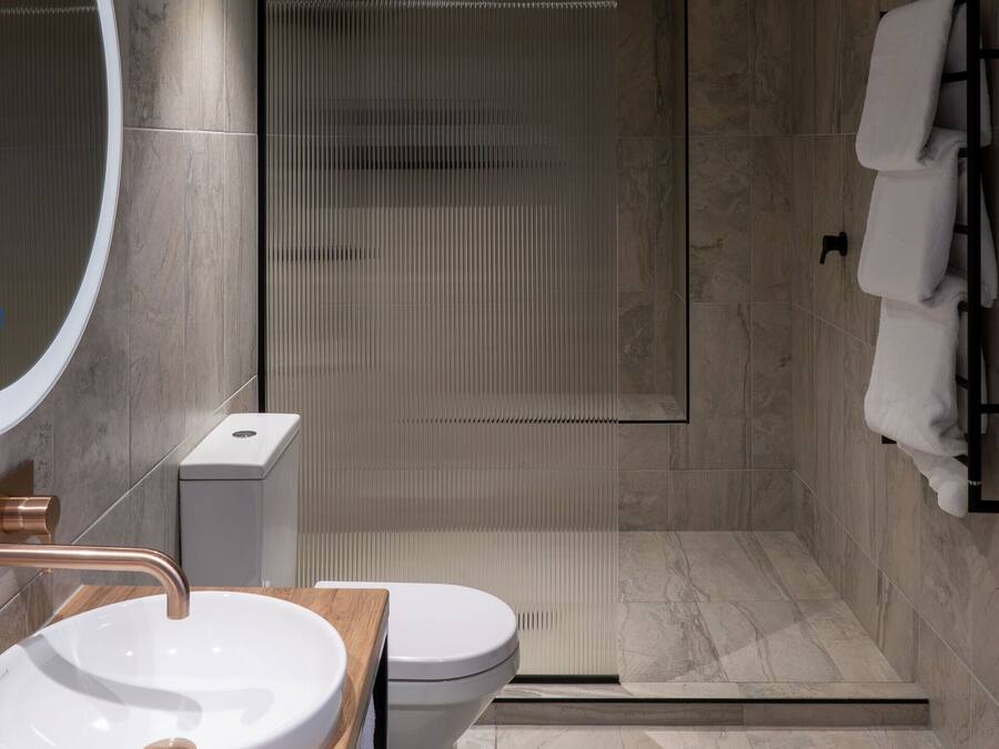 Une salle de bain moderne avec un meuble-lavabo en bois, un miroir rond, un lavabo blanc, des toilettes et un espace douche doté d’une porte en verre.