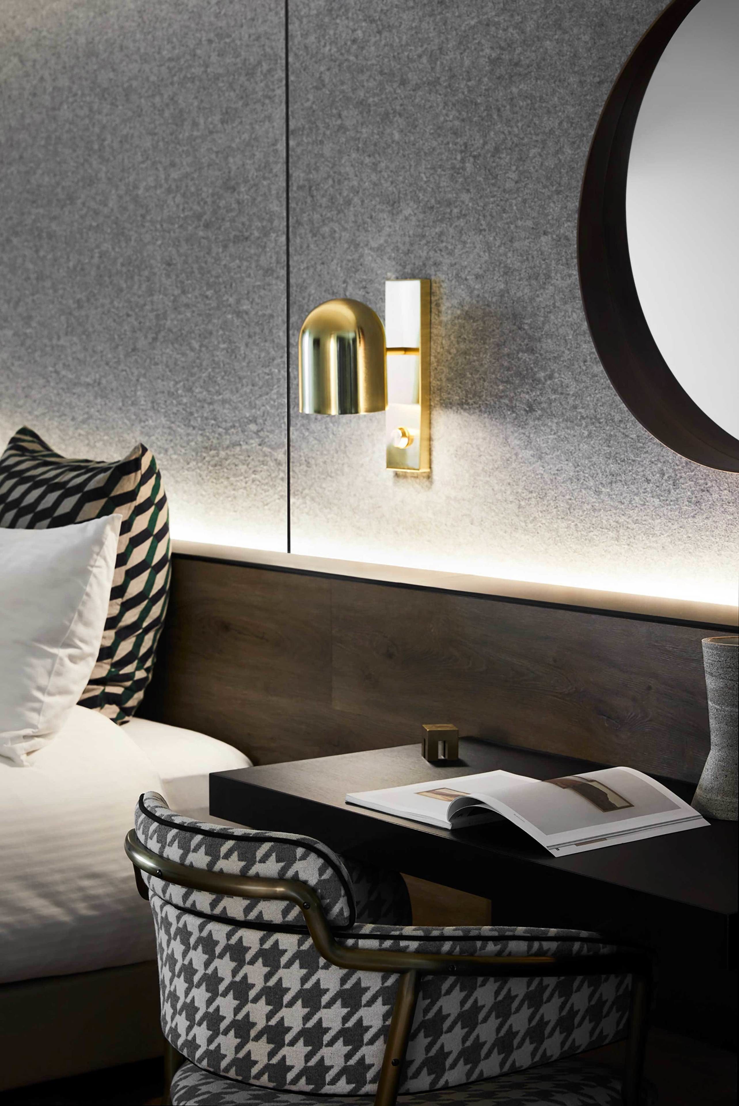 Ein modernes Hotelzimmer mit einem Ohrensessel im Hahnentrittmuster, einem Nachttisch mit einer Zeitschrift und einer Wandleuchte.