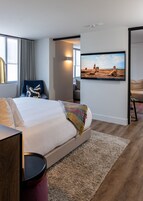Una habitación de hotel moderna con una cama grande, un televisor montado en la pared, una ventana con cortinas y vistas a un paisaje desértico.