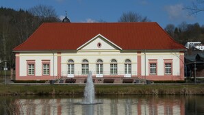 A building with a red roof and white walls, a fountain in front, and a pond reflecting the structure. - Hotel am Salzberg (Philippsthal)