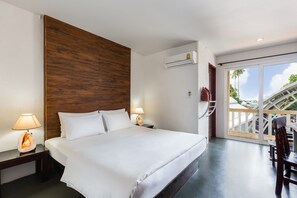 Une chambre d’hôtel avec un grand lit, une tête de lit en bois, deux lampes de chevet, un appareil de climatisation et un balcon offrant une vue sur les palmiers.