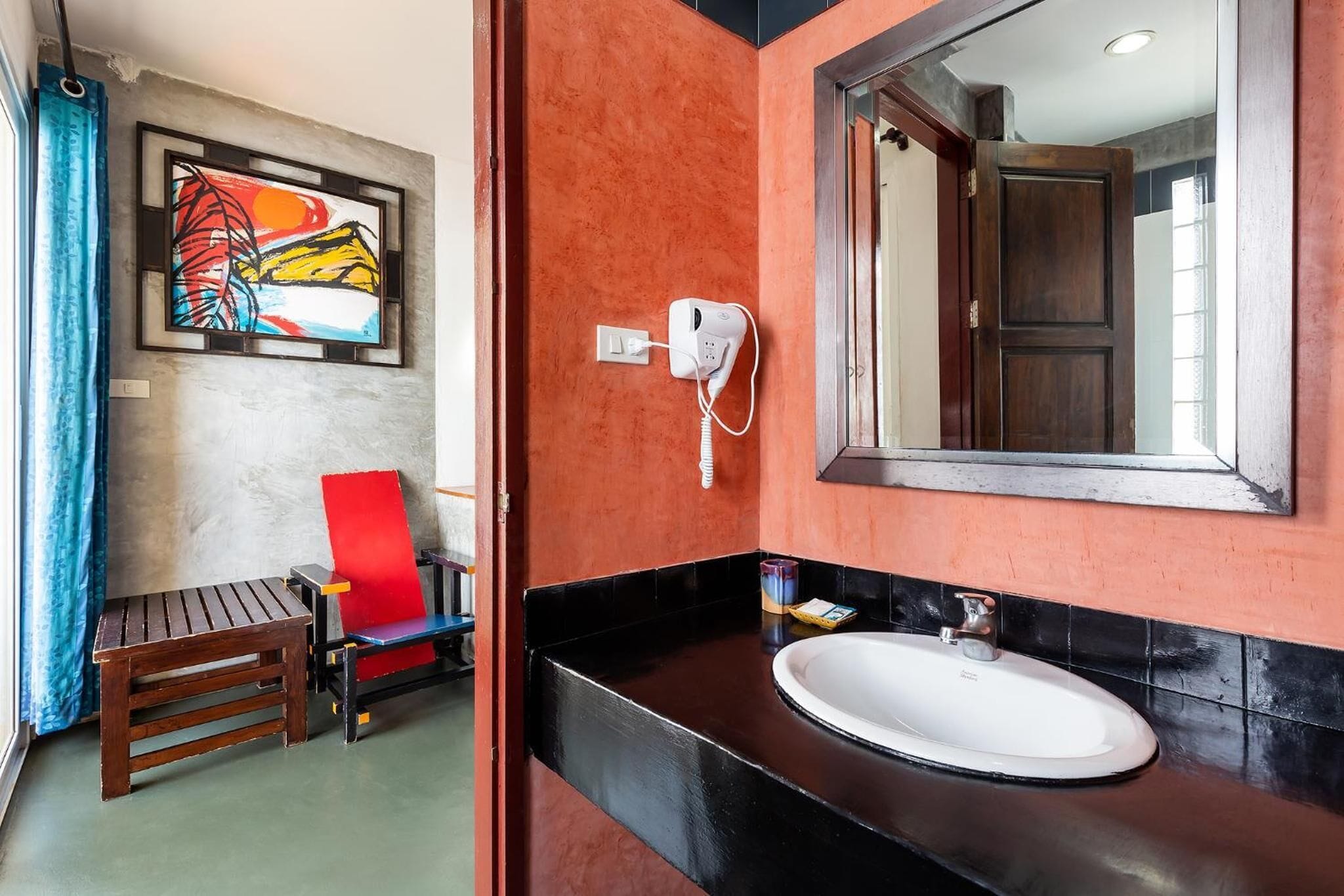 Ein Badezimmer mit roter Wand, schwarzem Waschtisch, weißem Waschbecken, einem roten Stuhl und einem gerahmten Kunstwerk.