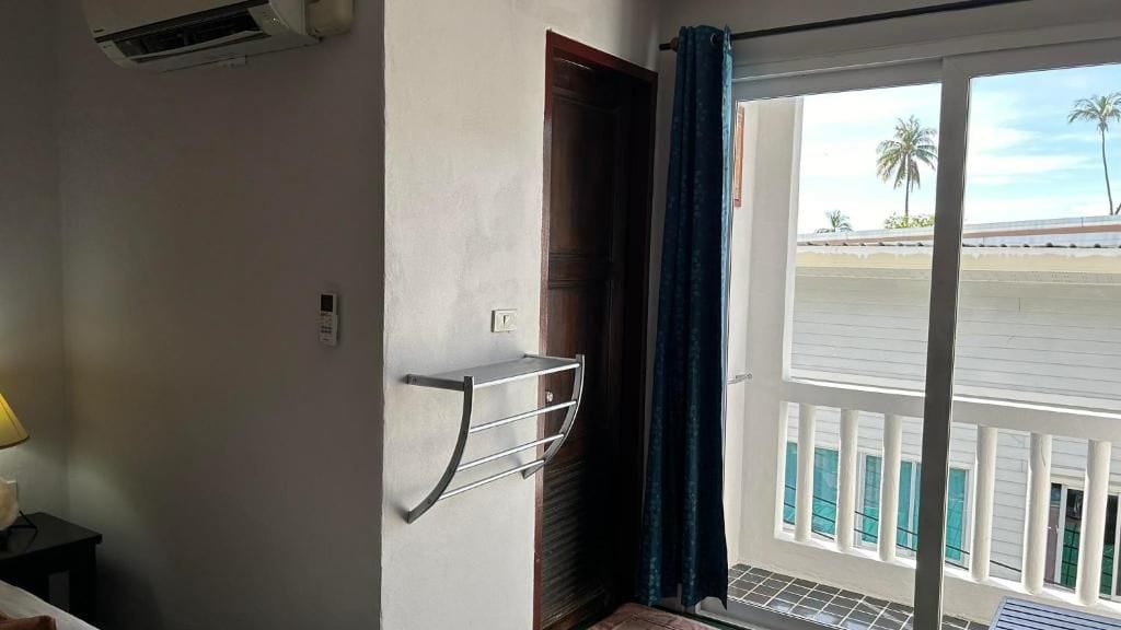 Ein Zimmer mit einem Bett, einer Wand-Klimaanlage, einem Fenster mit Blick auf Palmen und einem Balkon mit Geländer.