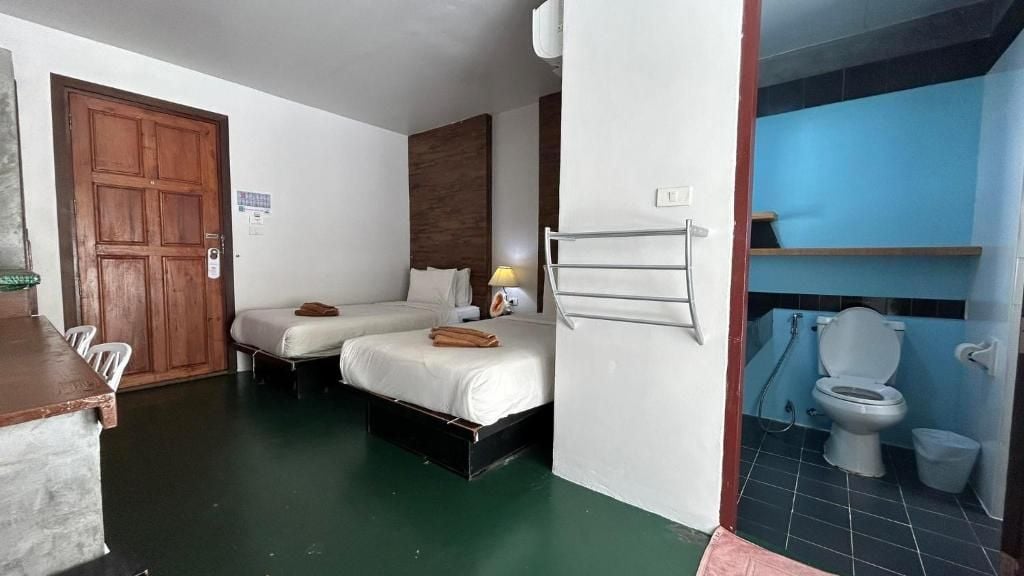 A hotel room with a wooden door, two beds, a small table, a chair, a towel rack, a nightstand, a lamp, and a bathroom with a toilet and sink.