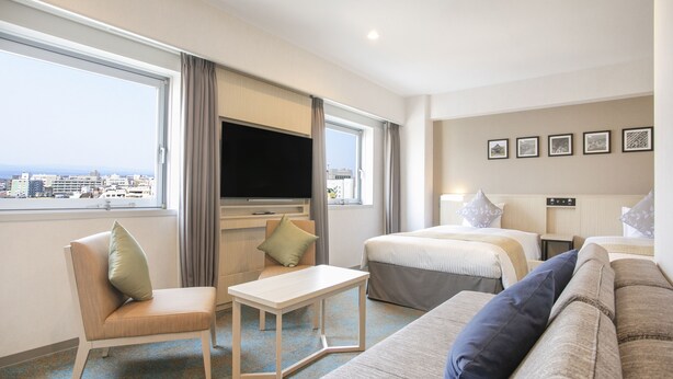A hotel room with a large window, a flat-screen TV, a sofa, a chair, a small table, and a bed. - Art Hotel Oita (Oita)