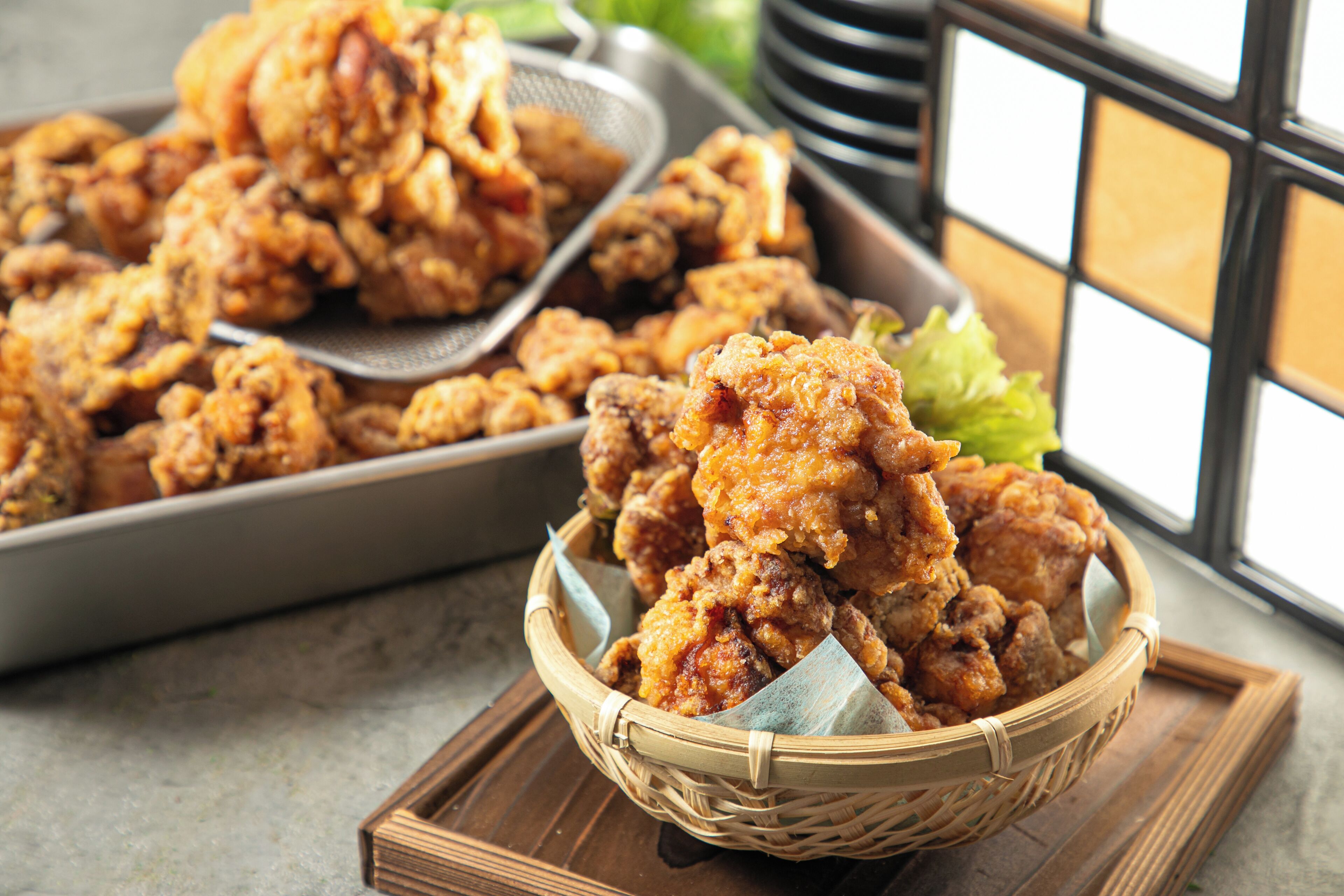 A basket of fried chicken with a metal strainer and a tray of food in the background.