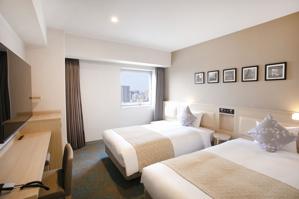 A hotel room with two beds, a desk, a TV, and a window with curtains.