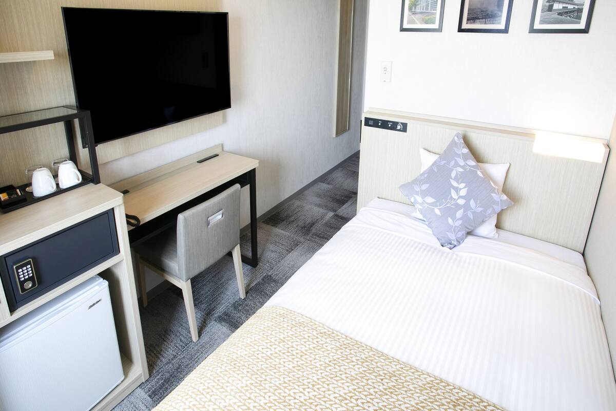 A hotel room with a bed, a TV, a desk with a computer, a mini-fridge, and a small safe.