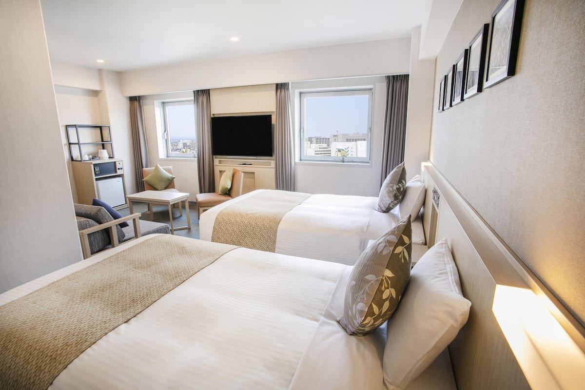 A hotel room with two beds, a TV, a desk, and a city view.