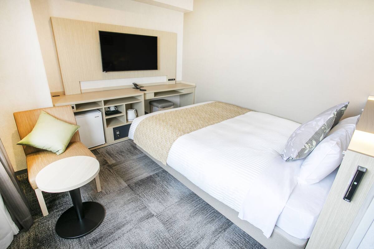 A hotel room with a bed, a desk, a chair, a TV, and a small table.