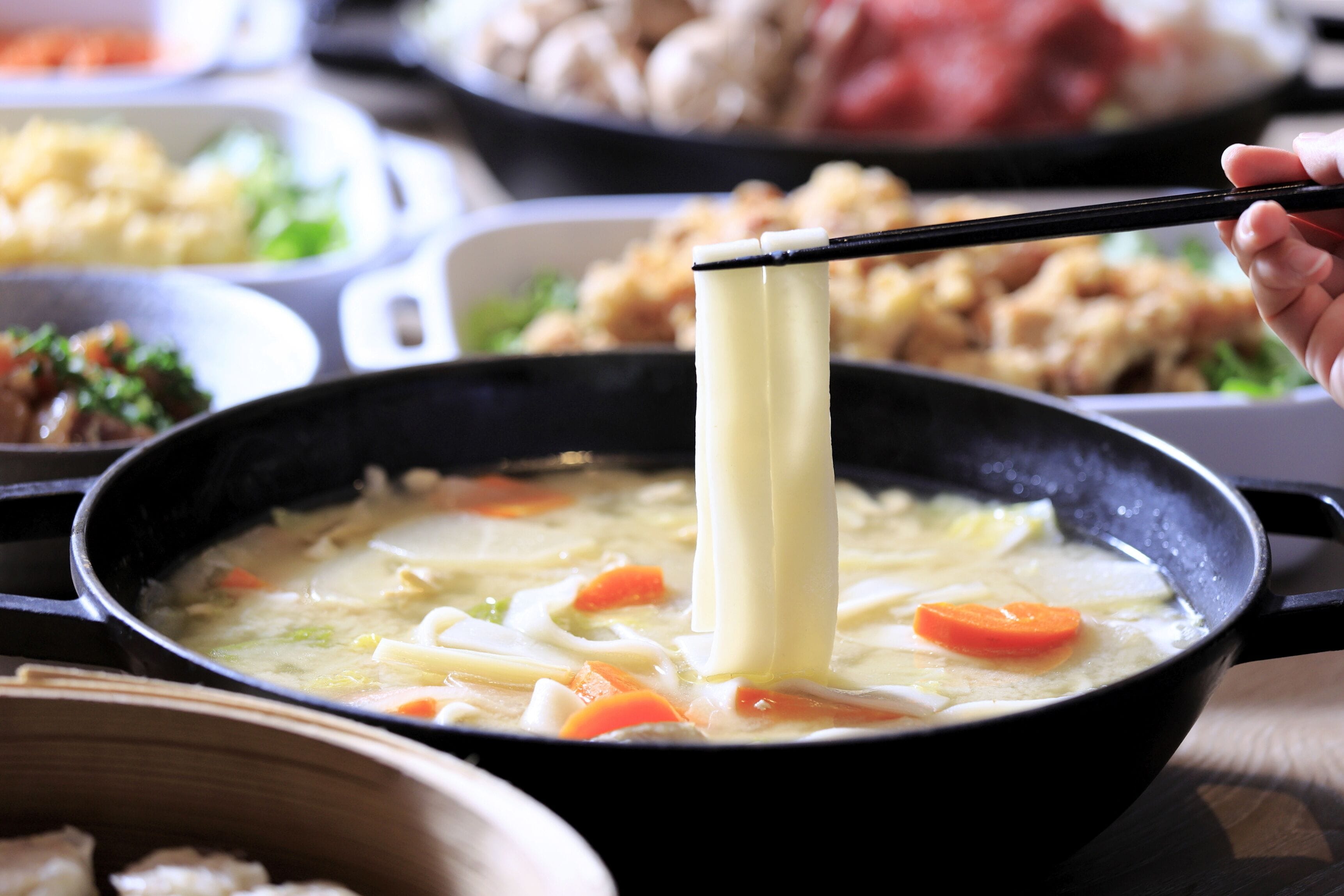 A person holding chopsticks over a pot of soup with noodles and vegetables.