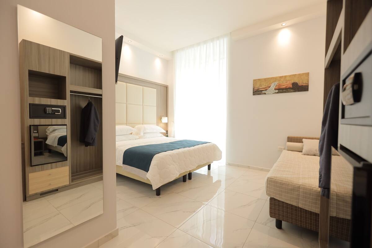 A modern hotel room with a large bed, a wardrobe with a built-in microwave, a small seating area, and a painting on the wall.