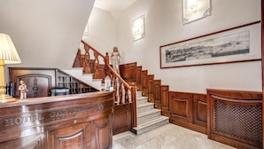 A hotel lobby with a wooden reception desk, a staircase with a wooden railing, and a framed historical map on the wall. - Garibaldi Suite (Naples)