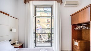 A hotel room with a bed, a wooden wardrobe, a nightstand, a window with a view of balconies, and an air conditioning unit. - Garibaldi Suite (Naples)