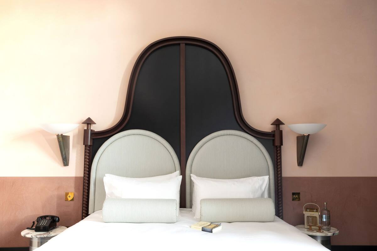 A hotel room with a large bed, two bedside lamps, a telephone, and a small table with a bottle and a book.