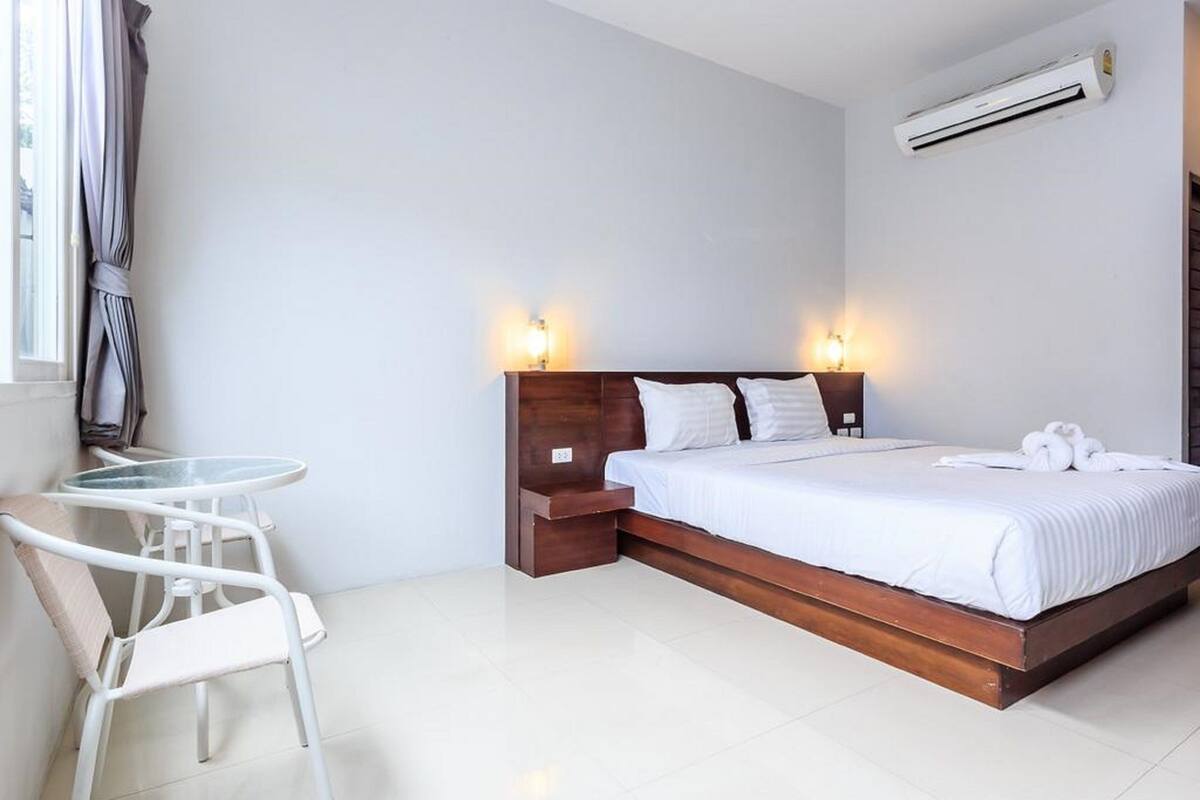 A hotel room with a bed, bedside tables, a small table with a chair, and an air conditioning unit.