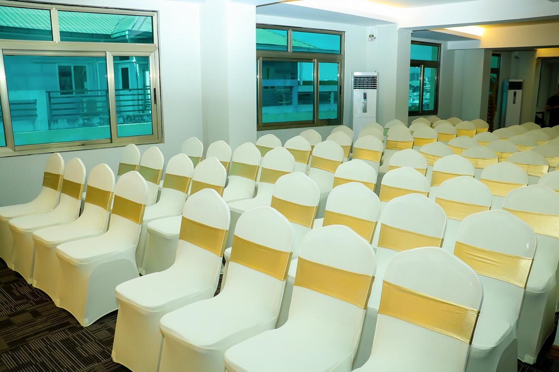 A conference room with rows of white chairs with gold sashes, large windows, and an air conditioning unit.