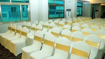 A conference room with rows of white chairs with gold sashes, large windows, and an air conditioning unit.