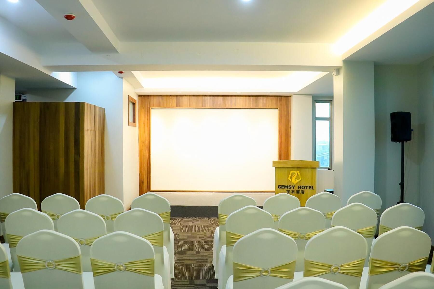Een vergaderzaal met rijen witte stoelen met gouden linten, een spreekgestoelte met de aanduiding 'GEMSY HOTEL' en een groot leeg scherm aan de voorzijde.