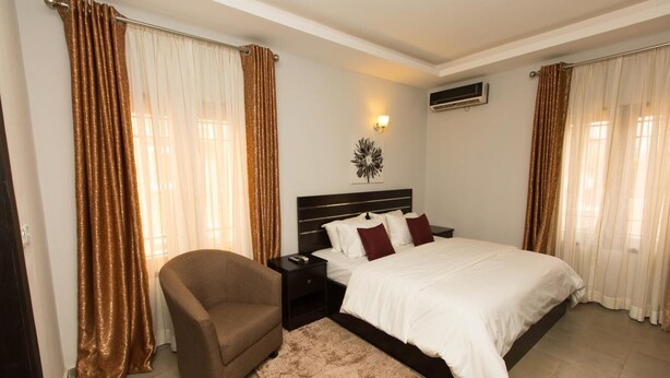 A bedroom with a bed, nightstand, chair, and curtains. - Vulcan Suites (Abuja)