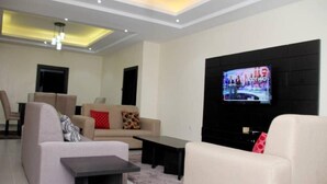 A modern living room with a flat-screen TV mounted on a black wall, a beige sofa, a coffee table, and a light-colored armchair. - Vulcan Suites (Abuja)