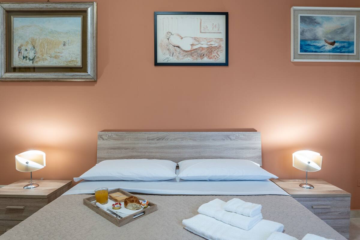 A neatly arranged hotel room with a bed, two bedside lamps, a tray with breakfast items, and artwork on the walls.