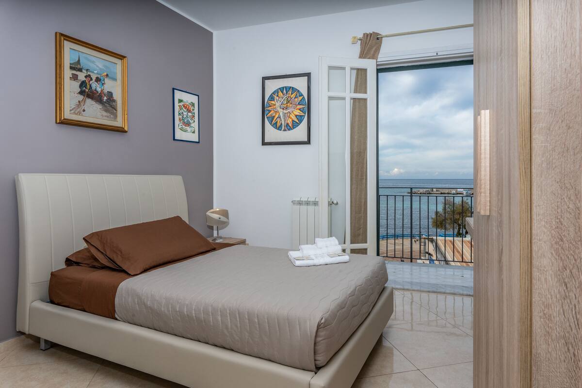 A bedroom with a bed, a nightstand, a window with a view of the sea, and two framed pictures on the wall.