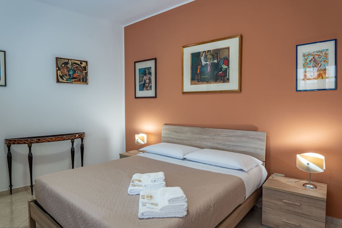 A neatly made bed with white linens and towels, a bedside table with a lamp, and framed artwork on the walls.