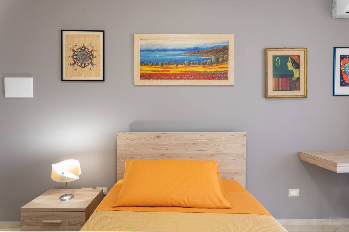 A bedroom with a wooden headboard, an orange pillow, a bedside lamp, and framed artwork on the wall.