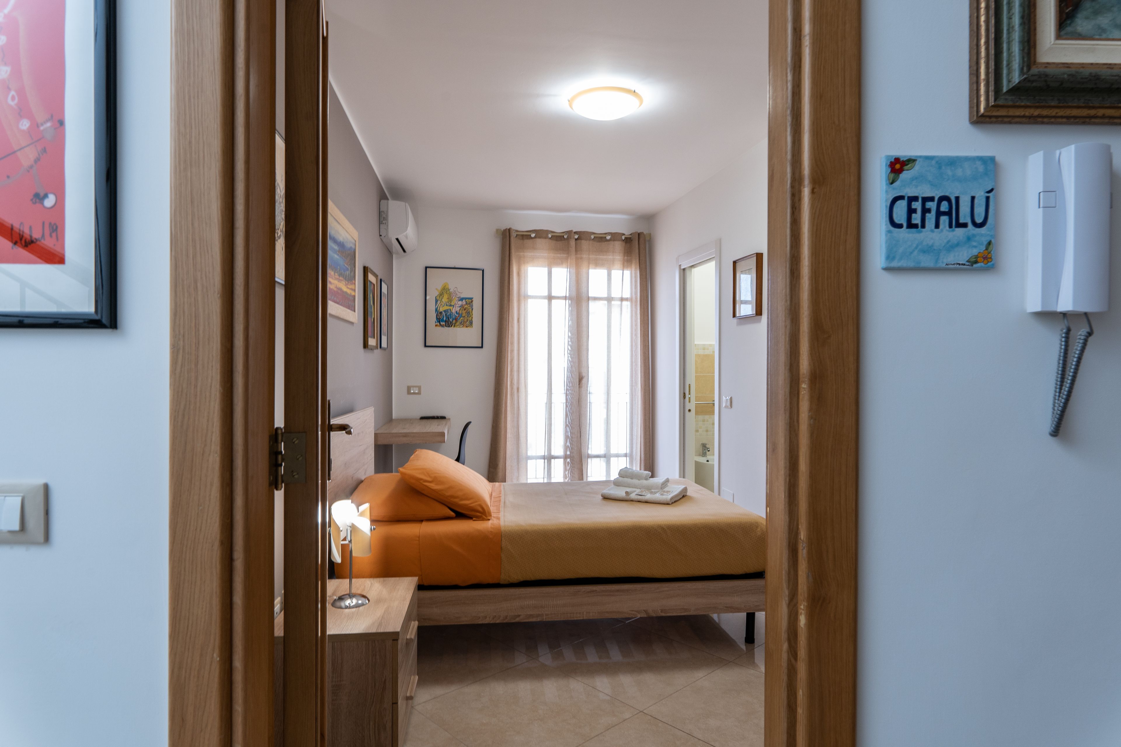 A bedroom with a bed, a bedside table, a window with curtains, and a wall-mounted phone.