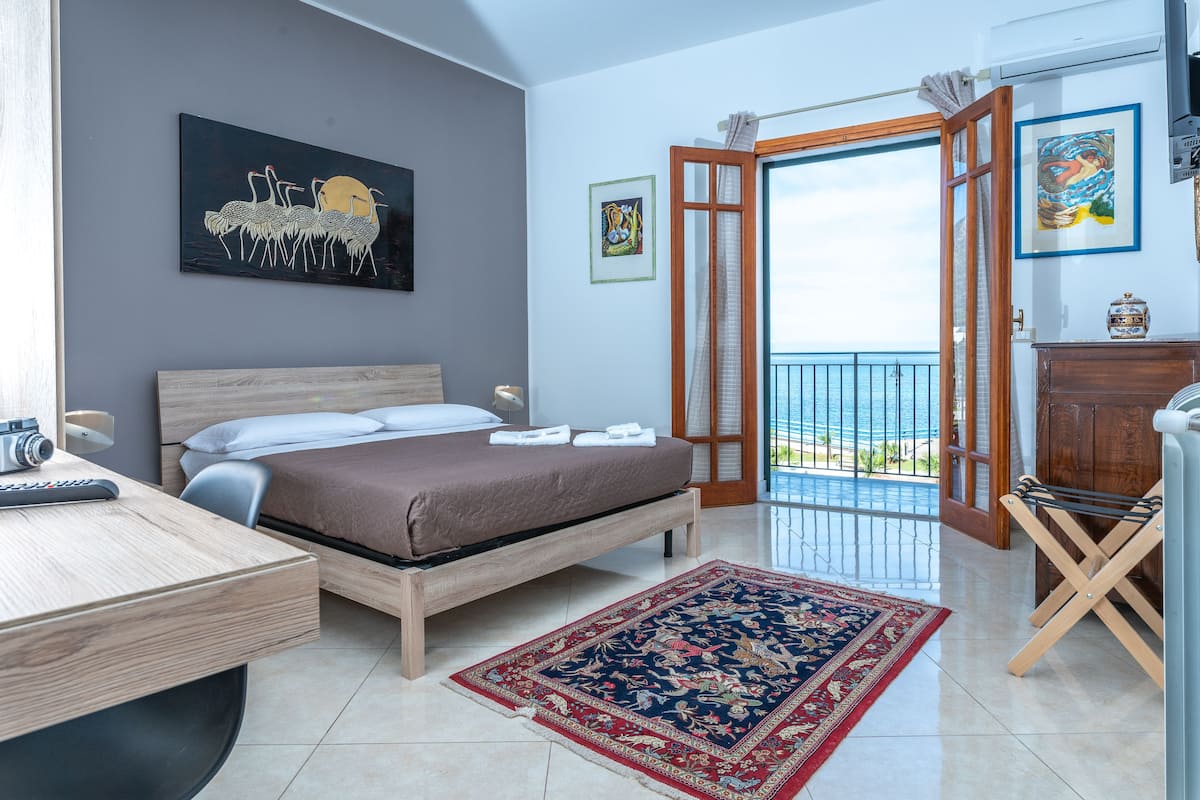 A bedroom with a bed, a desk, a chair, a balcony with a view of the sea, and artwork on the walls.