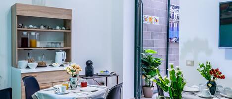 A modern dining area with a table set for four, a wooden shelf with kitchenware, and a potted plant.