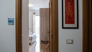 A hallway with a wooden door leading to a bedroom with a bed and a window. There is a framed picture on the wall and a small sign with the word "ERICE". - Una Terrazza sul Golfo (Palermo)