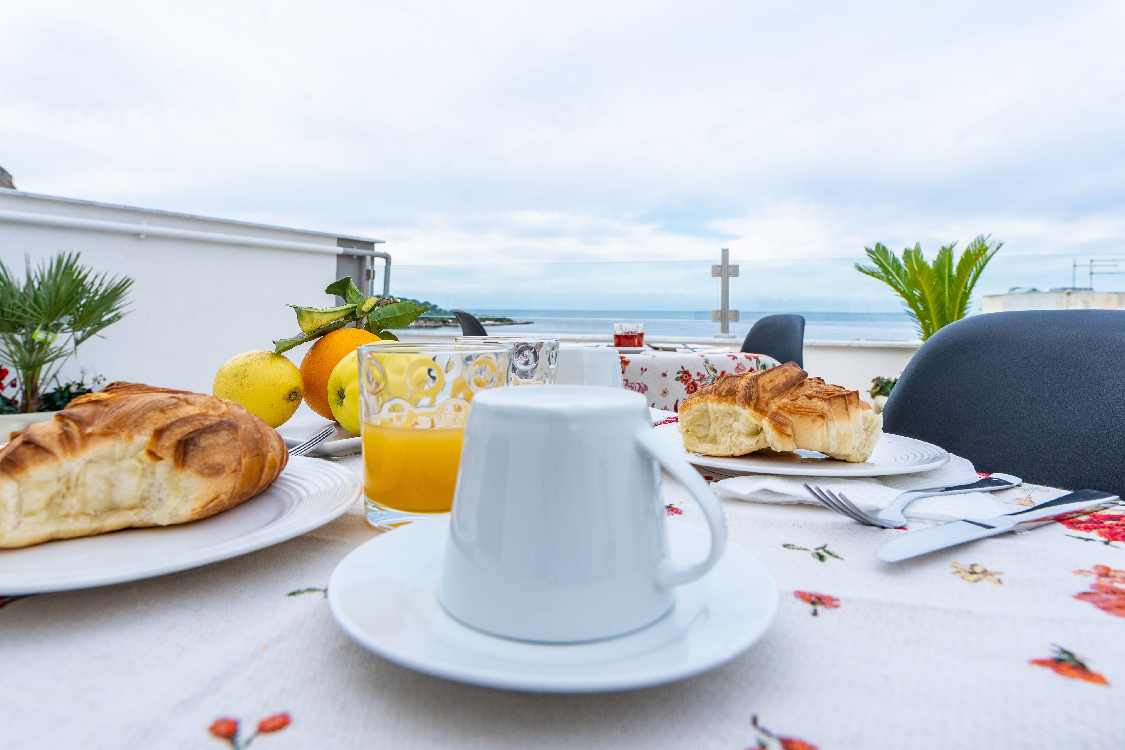 Ein Frühstücksbuffet mit Croissants, Orangensaft und einer Tasse auf einem Tisch mit einer blumigen Tischdecke, mit Blick auf ein Gewässer und ein Kreuz.