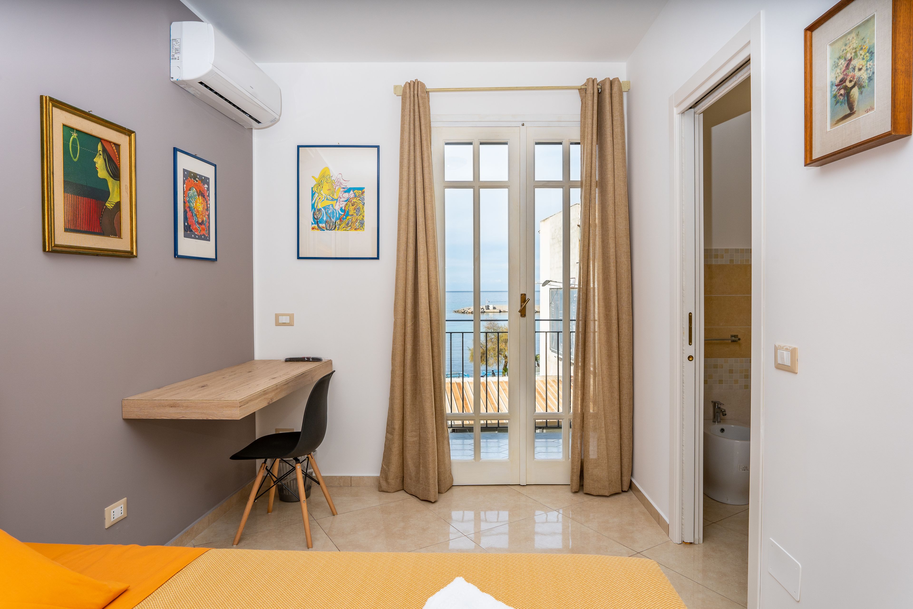 Una habitación con escritorio de madera, una silla, una cama con manta naranja y un balcón con cortinas.