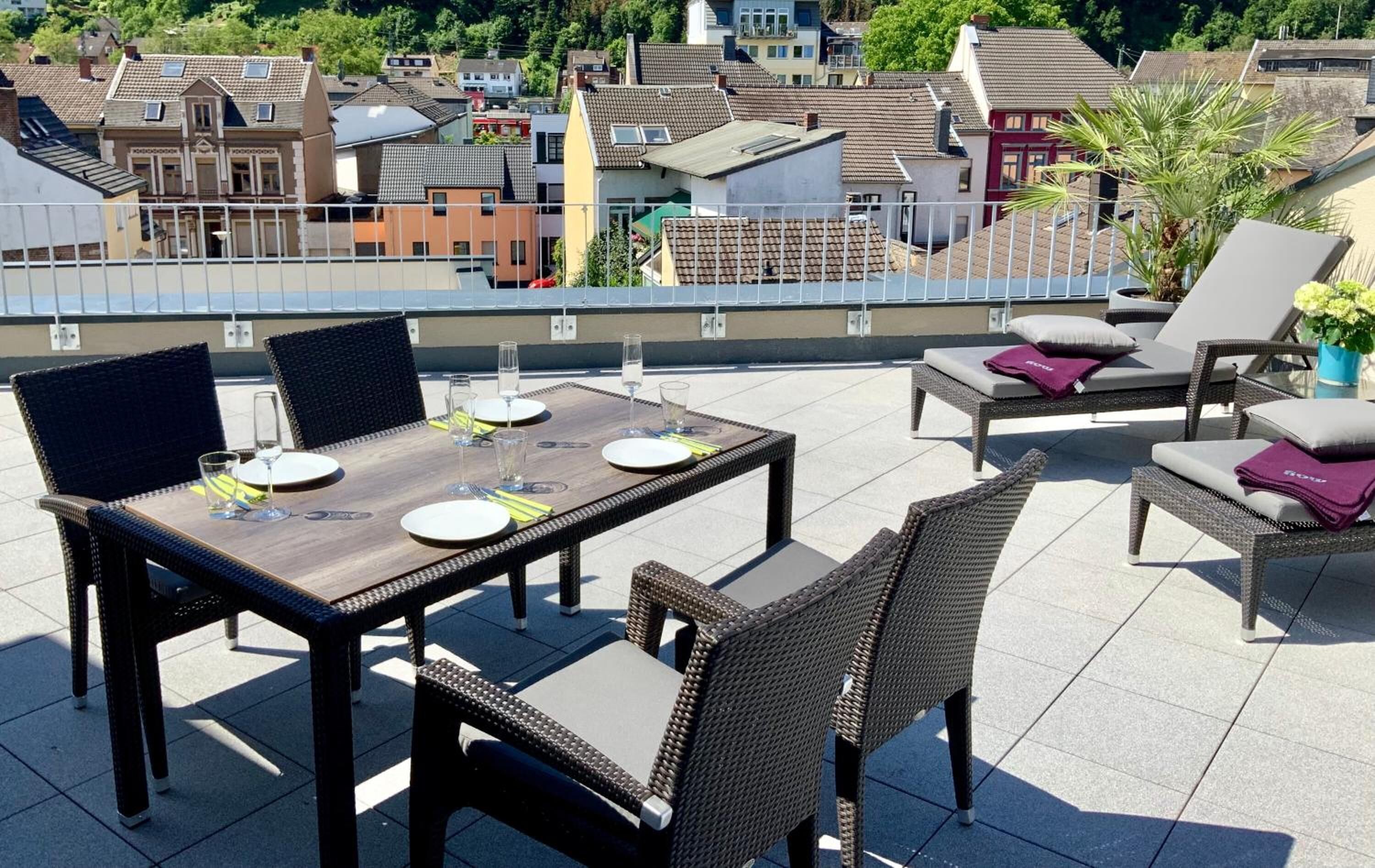 A rooftop terrace with a dining table and chairs, a lounge area with a sofa and armchairs, and a view of the cityscape in the background.