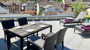 A rooftop terrace with a dining table and chairs, a lounge area with a sofa and armchairs, and a view of the cityscape in the background.